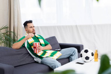 Çizgili eşarplı spor fanatiği futbol topunun yanında televizyonda maç izliyor.