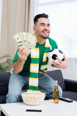 happy bookmaker holding dollars and soccer ball while watching match on tv