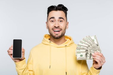 cheerful bookmaker holding dollar banknotes and smartphone with blank screen isolated on grey