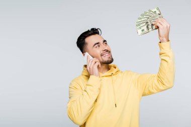pleased bookmaker holding dollar banknotes while talking on cellphone isolated on grey