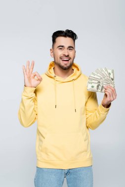 excited bookmaker holding dollars and showing okay sign isolated on grey clipart