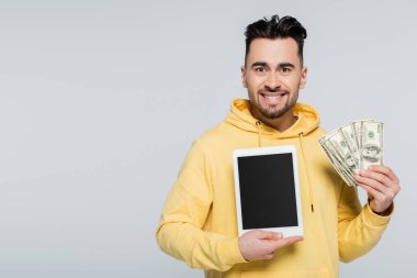 joyful gambler holding dollars and digital tablet with blank screen isolated on grey
