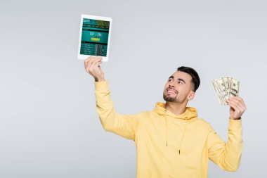 KYIV, UKRAINE - 4 Şubat 2022: Gri üzerine izole edilmiş spor uygulaması ile dolar ve dijital tablet tutan neşeli bahisçi