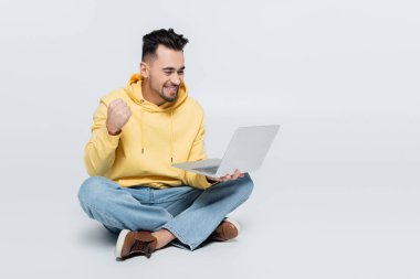 excited bookmaker showing success gesture near laptop while sitting on grey