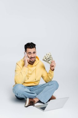 happy bookmaker with money sitting near laptop and talking on mobile phone on grey