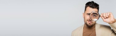young skeptical critic looking at camera through magnifier glass isolated on grey, banner