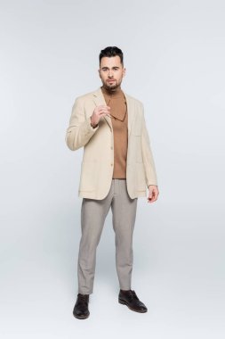full length of critic in beige blazer and trousers holding eyeglasses on grey