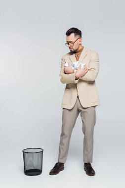 full length view of critic with crumpled paper standing near trash can on grey