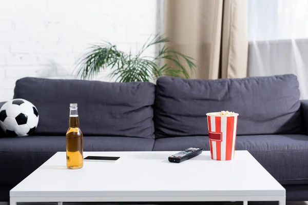 テーブルにはビールとテレビのリモコンとポップコーンのバケツとソファの上のサッカーボールの近くのスマートフォン — ストック写真