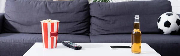 sofa with soccer ball near table with bucket of popcorn, beer, tv remote controller and smartphone, banner