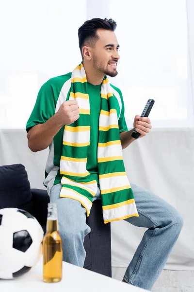 家でサッカーの試合を見ながら幸運のために拳を握りしめている縞模様のスカーフで興奮した男 — ストック写真