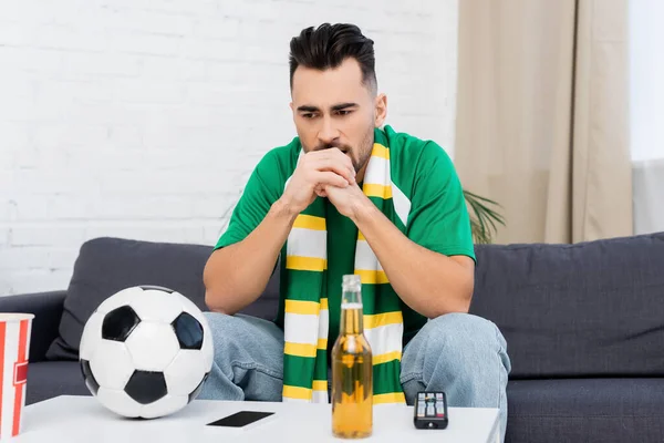 frustrated sports fan sitting near tv remote controller, soccer ball and beer