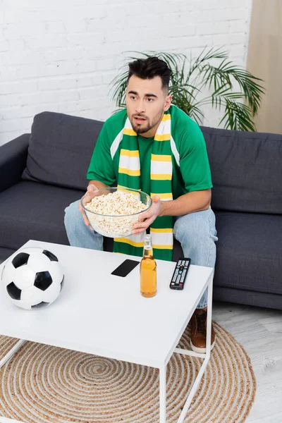 surprised sports fan with bowl of popcorn watching match on tv near beer and soccer ball