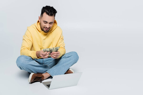 pleased man counting dollars while sitting with laptop on grey