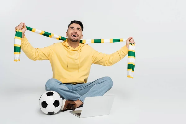 joyful sports fan holding striped scarf while sitting near laptop and soccer ball on grey