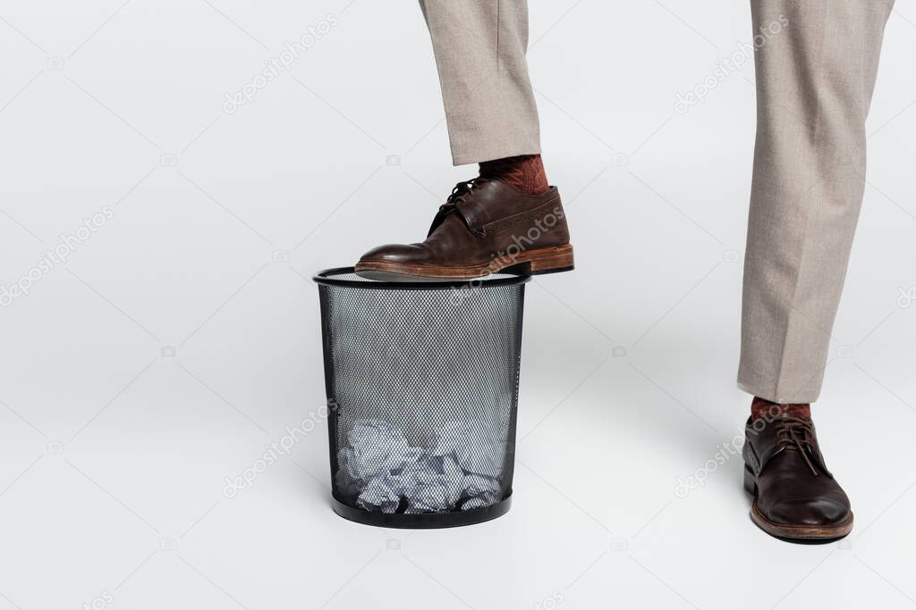 cropped view of critic stepping on trash can with crumpled paper on grey