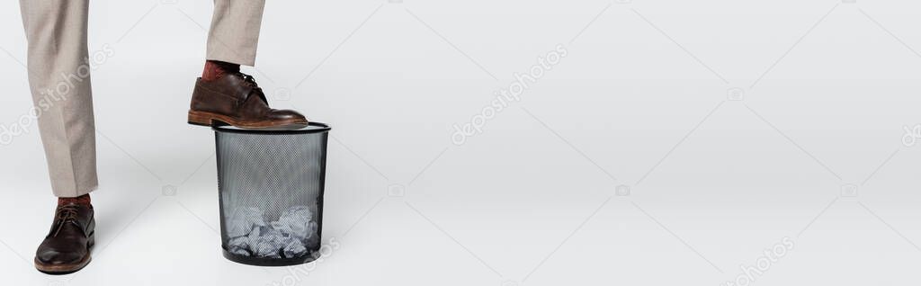 partial view of man stepping on trash can with crumpled paper on grey, banner