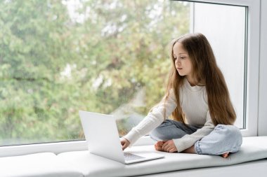 Kız bacak bacak üstüne atmış pencere kenarında oturuyor ve online ders sırasında dizüstü bilgisayar kullanıyor.