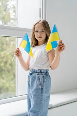 Küçük Ukrayna bayraklı vatansever kız pencerenin yanında durup kameraya bakıyor.
