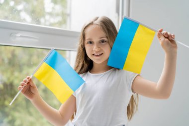 Küçük Ukrayna bayraklı kız evinin penceresinin yanında gülümsüyor.