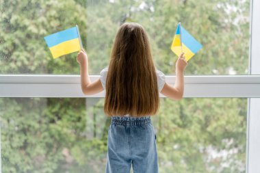 Pencerenin yanında küçük Ukrayna bayraklarıyla ayakta duran uzun saçlı kızın arkadan görünüşü