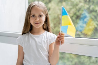 Küçük Ukrayna bayrağı taşıyan mutlu kız evdeki pencerenin yanındaki kameraya bakıyor.