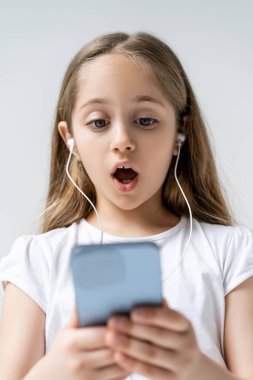 Kulaklıklı, gri renkte izole edilmiş, bulanık akıllı telefona bakan hayrete düşmüş bir kız.