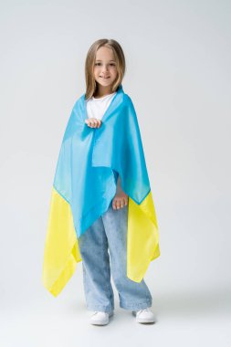 full length view of positive girl covered with ukrainian flag on grey clipart