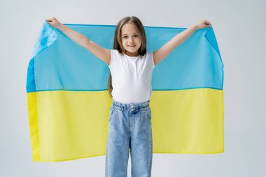 Beyaz tişörtlü ve kot pantolonlu mutlu kız gri üzerinde Ukrayna bayrağı taşıyor.