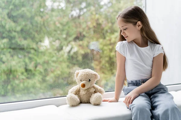 穿着牛仔裤的女孩坐在窗台上 靠近泰迪熊 戴着雨滴玻璃杯 — 图库照片