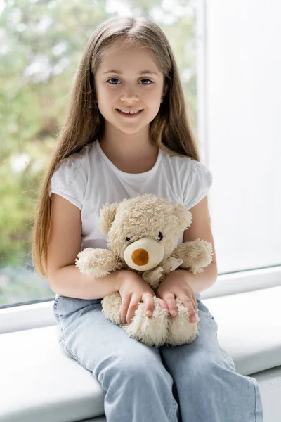 Veselá Dívka Usmívá Kameru Zatímco Sedí Okna Plyšovým Medvídkem — Stock fotografie