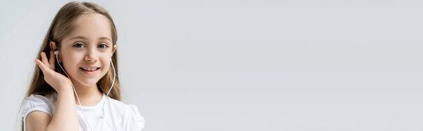 smiling girl adjusting earphone and smiling at camera isolated on grey, banner