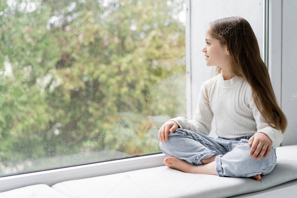 barefoot girl in jeans sitting on windowsill with crossed legs and looking through glass with raindrops