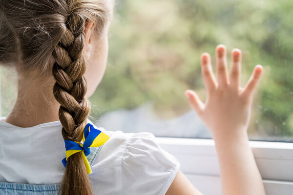 back view of girl with blue and yellow ribbon on braid standing near blurred window