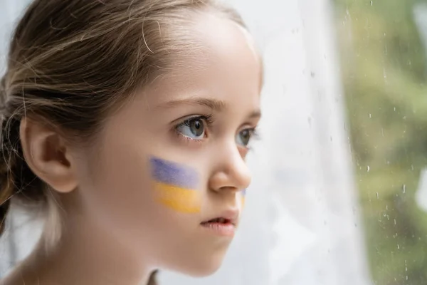 portrait of girl with ukrainian flag on face looking away through window with raindrops