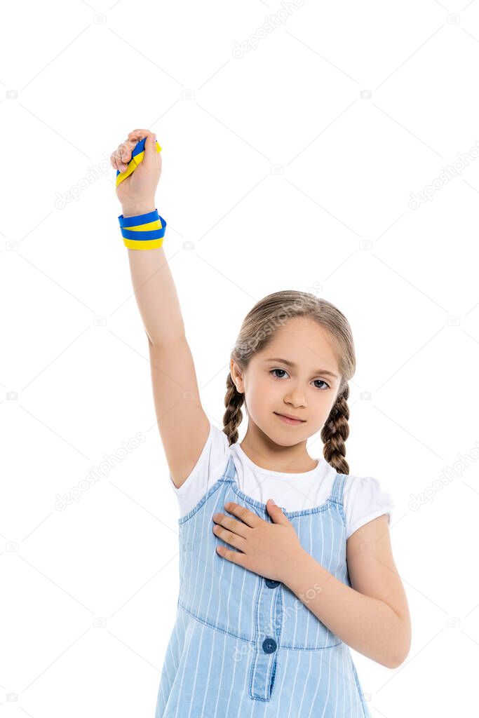 patriotic ukrainian girl with blue and yellow ribbon touching heart while standing with raised hand isolated on white