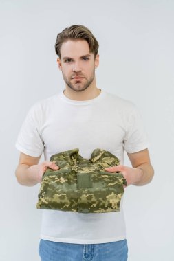 Genç adam askeri üniforma giyiyor ve beyaz kameraya bakıyor. 
