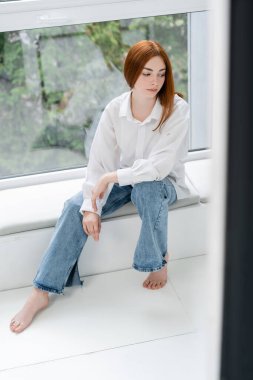 Kotlu ve gömlekli kızıl saçlı kadın pencere eşiğinde oturuyor. 