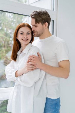 Genç adam, evin penceresinin yanında tişörtlü neşeli kız arkadaşına sarılıyor. 