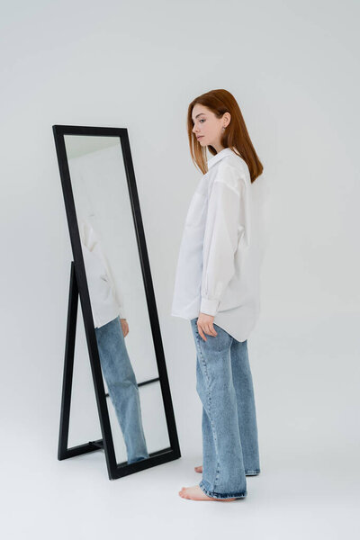 Side view of red haired woman standing near mirror on white background