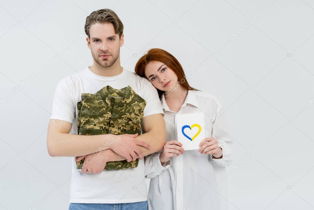 Young man holding military uniform near girlfriend with blue and yellow heart on card isolated on white 