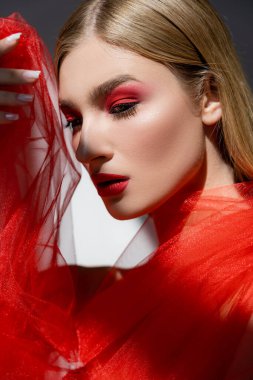 Kırmızı dudaklı genç bir kadın gri arka planda tül kumaş tutuyor. 