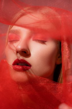 Kırmızı makyajlı, kapalı gözlü, gri arkaplanda bulanık bir kumaş gibi duran genç bir kadın. 