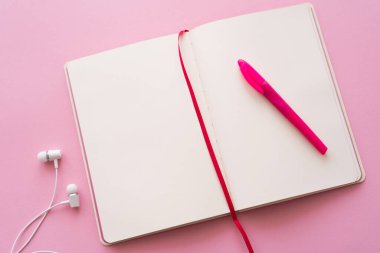 Pembe üzerine kablolu kulaklıkların yanındaki açık defter ve kalemin üst görünümü 