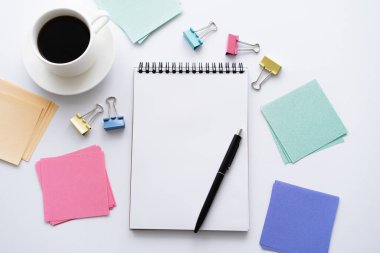 top view of pen on notebook near paper notes, fold back clips and cup of coffee on white