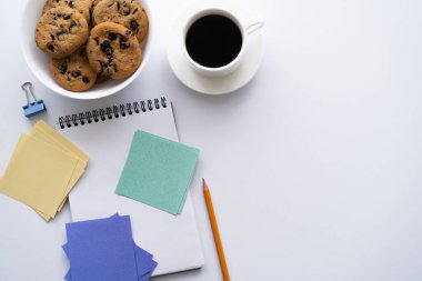 top view of bowl with chocolate chip cookies near cup of coffee and stationery on white