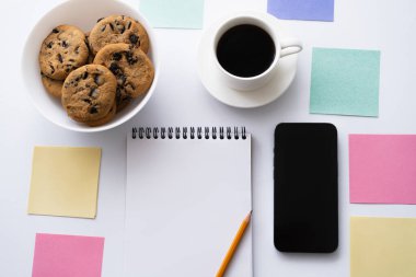 top view of cup of coffee, cookies and stationery near smartphone with blank screen on white
