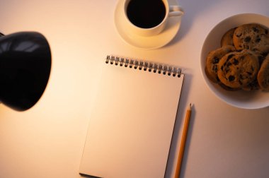 top view of lamp near chocolate chip cookies, cup of coffee and notebook on white
