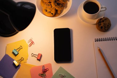Beyaz ekranlı akıllı telefonun yanındaki lambanın üst görüntüsü, çikolatalı kurabiyeler, beyaz üzerine bir fincan kahve ve kırtasiye.