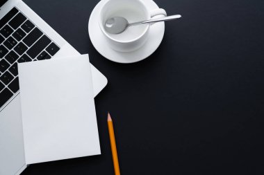 top view of blank paper on laptop near pencil and empty cup with spoon on black 